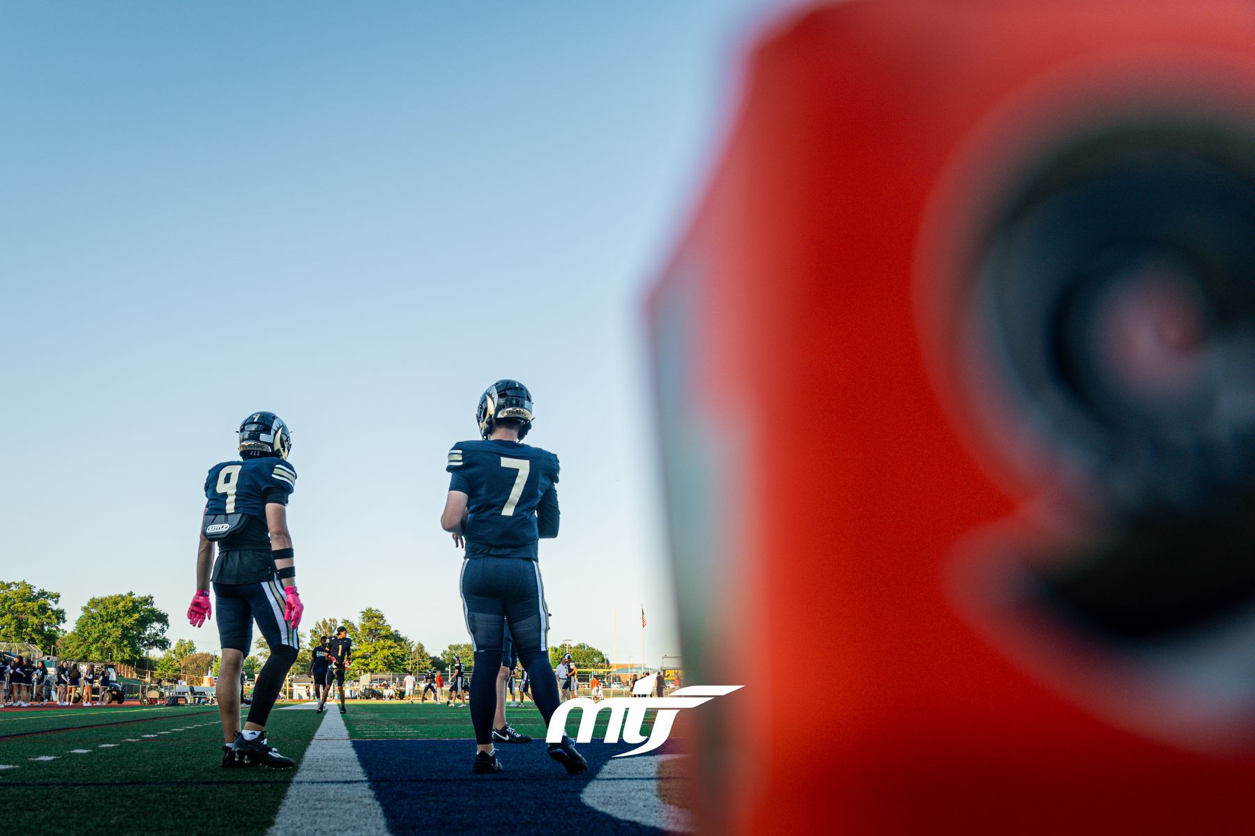 Two football players waiting on the sideline at sunset, an end-zone pylon blurred in the foreground